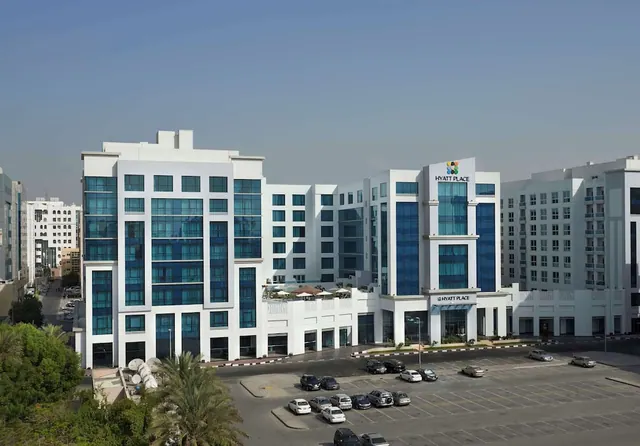 Exterior view of a white multi-storey Hyatt Place hotel with blue glass windows, a parking lot in front and palm trees at the left edge.