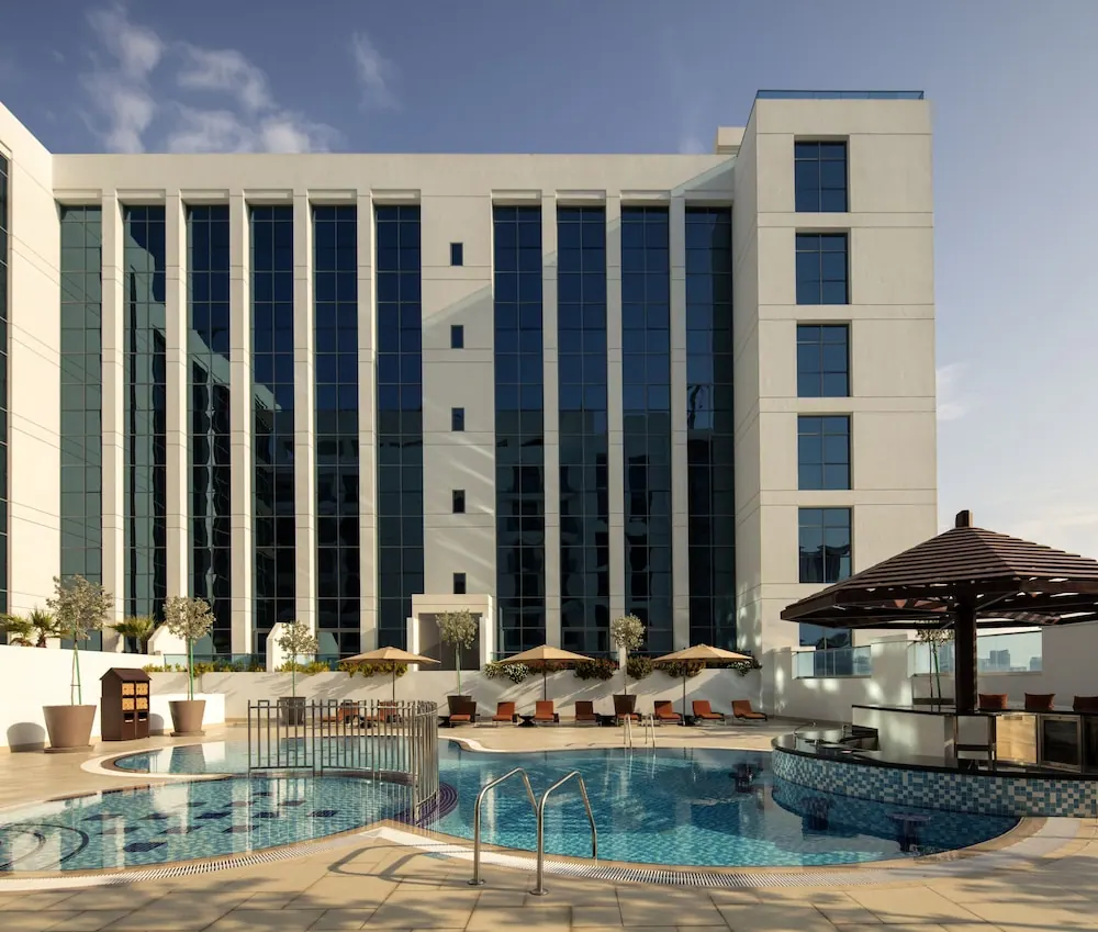 An outdoor pool with orange sun loungers and parasols sits in front of a white multi-storey hotel building, with a wooden pergola and bar counter on the right.