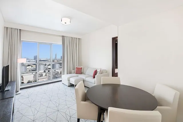 A living and dining area with a light grey sofa, a round dark dining table with cream chairs, and floor-to-ceiling windows looking out over city buildings.