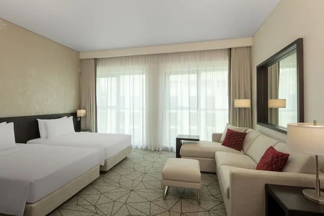 A hotel room with two beds with white bedding, a beige sofa with red cushions, an ottoman, and sheer-curtained floor-to-ceiling windows.