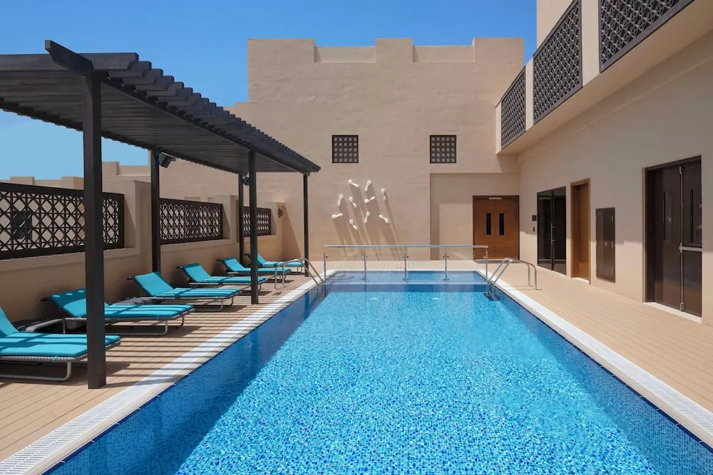 A rectangular outdoor swimming pool with teal sun loungers under a wooden pergola, beige walls with latticed screens, and a clear blue sky.