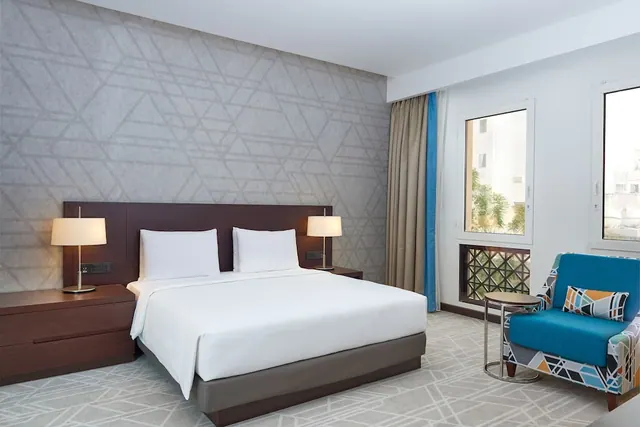 A made-up king bed with two bedside lamps and a patterned blue armchair sits in a hotel room, with a window showing trees and buildings outside.