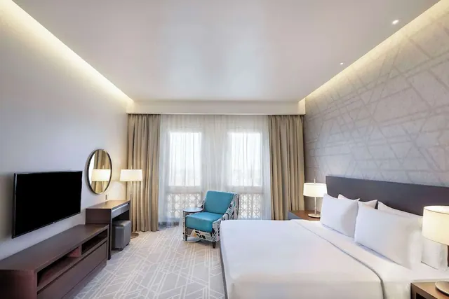 A hotel bedroom with a large white bed, a blue armchair by the sheer-curtained window, a wall-mounted television and a round mirror above a desk.