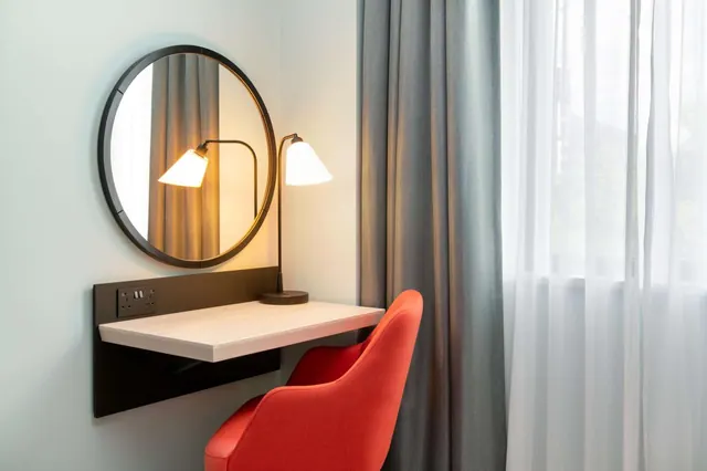 A wall-mounted desk with a round mirror, a table lamp and a red chair sits beside grey curtains and a sheer-curtained window in a bedroom.