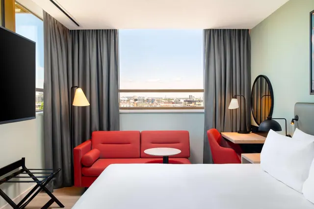 A hotel bedroom with a white double bed, a red sofa and round table, a desk with a lamp, and a window looking out over a city skyline.