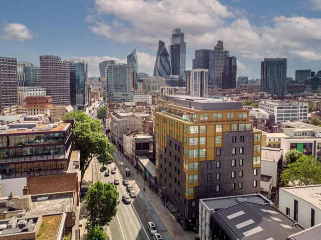 Elevated view over a city street and rooftops toward a cluster of tall office towers, including the Gherkin, under a partly cloudy sky.
