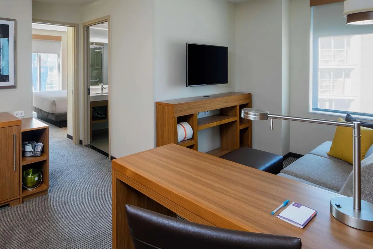 A hotel suite living area with a wooden desk and lamp, a wall-mounted television, a grey sofa by the window, and a view through the doorway to the bedroom and bathroom.