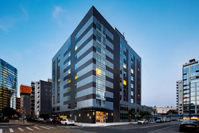 A multi-storey modern building with dark and light striped cladding and lit windows stands at a street corner at dusk, with parked cars along the road.