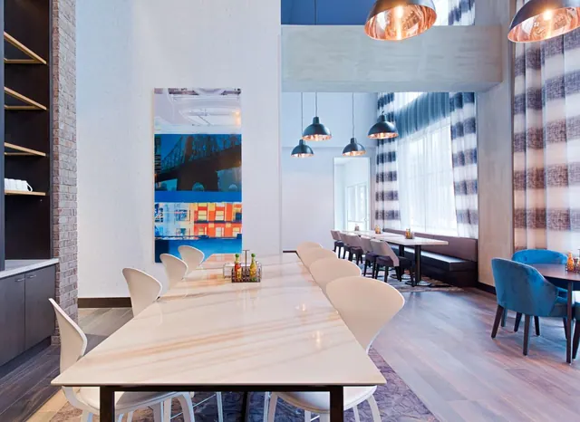 A dining room with a long marble-topped table, white chairs, copper pendant lamps, a banquette along the wall and a large blue artwork on the wall.