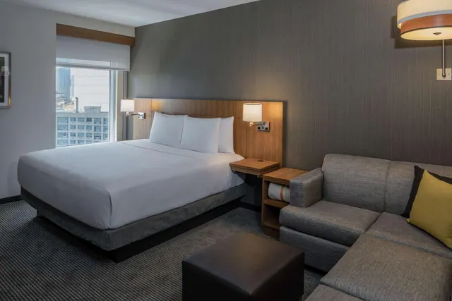 A king bed with white linens sits beside a grey sectional sofa, with a window showing city buildings outside.