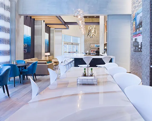 A hotel lobby with a long white communal table, white chairs, blue upholstered armchairs and sofas, and a reception counter in the background.