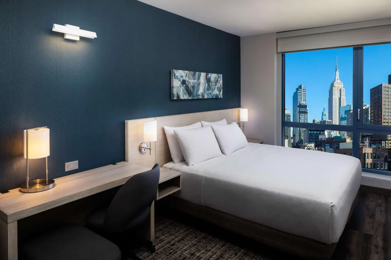 A hotel bedroom with a king bed, a desk and chair, and a large window showing the Empire State Building and surrounding city buildings.