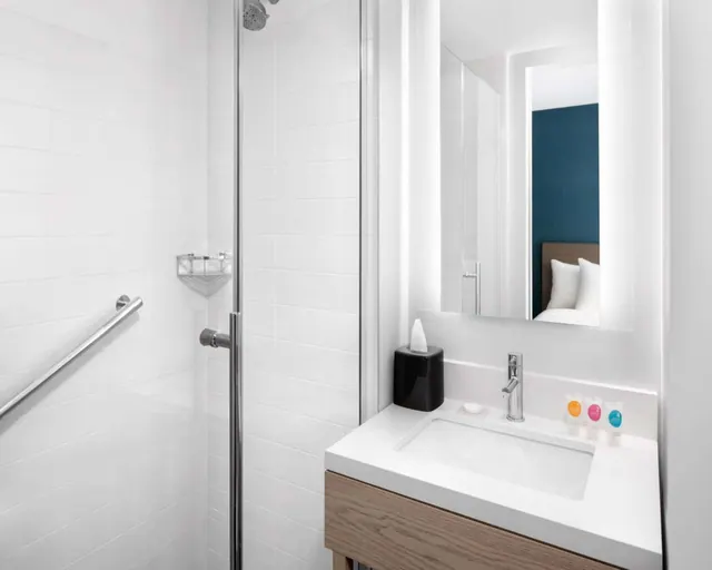 A bathroom with white tiled walls, a glass-enclosed walk-in shower with a grab rail, and a wood-base vanity with a sink, with a bedroom reflected in the mirror.