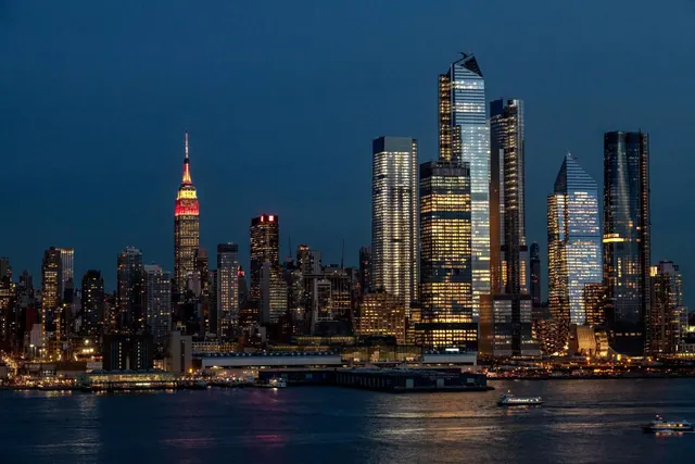 Dusk view of the Manhattan skyline across the Hudson River, with the Empire State Building lit in red and gold on the left and boats on the water in front.