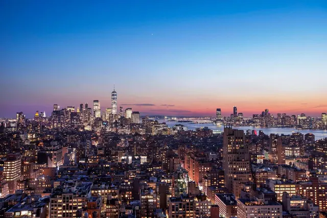 Aerial view at dusk over a dense city grid, with the One World Trade Center tower and the Hudson River visible beyond.