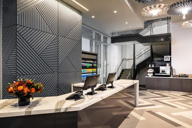 A hotel lobby reception desk with two monitors, telephones and a flower arrangement, with a staircase and coffee station in the background.