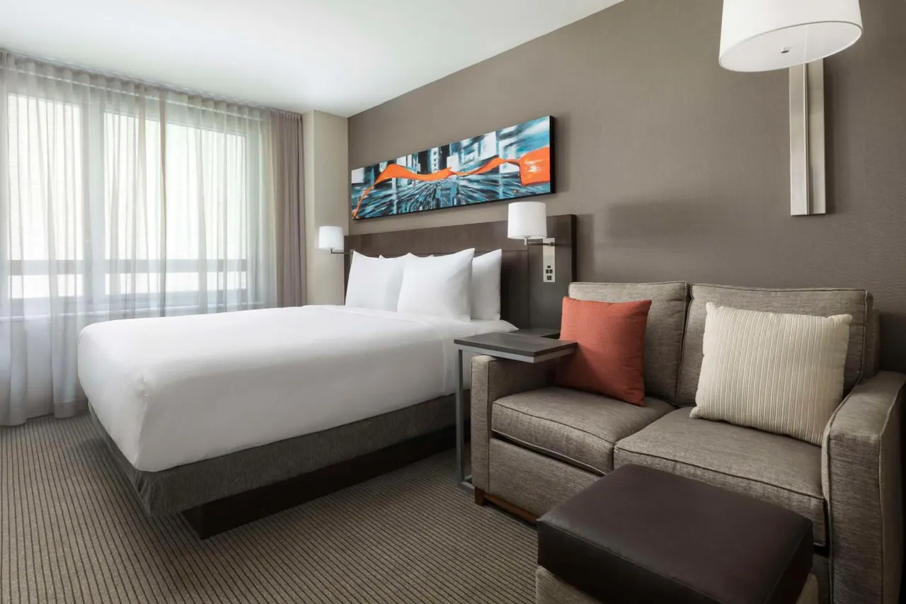 A king bed with white linens sits beside a grey sofa with orange and cream cushions, below a wall artwork, with a large window covered by sheer curtains on the left.