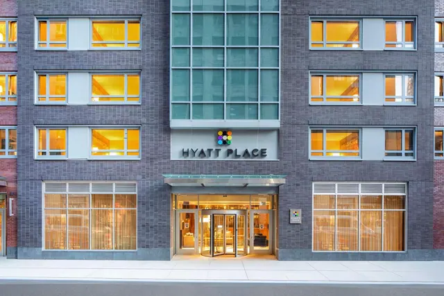 The front facade of a Hyatt Place hotel building at dusk, with lit windows, a central entrance canopy, revolving doors and the hotel name on the sign above the entrance.
