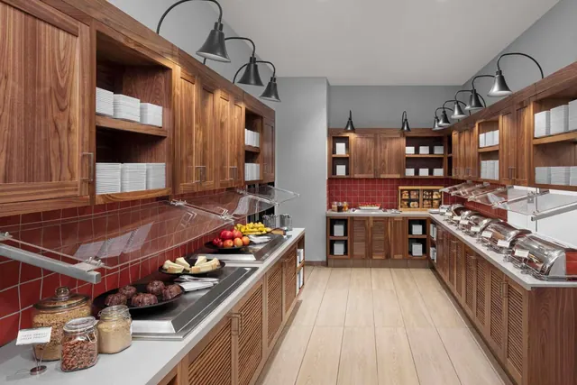 A breakfast buffet counter with wooden cabinets, a red tiled backsplash, sneeze guards, fruit, pastries and chafing dishes, with no people present.