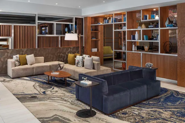 A lobby seating area with navy and beige sofas, low wooden coffee tables, a floor lamp and wood-fronted shelving holding books and decorative objects.