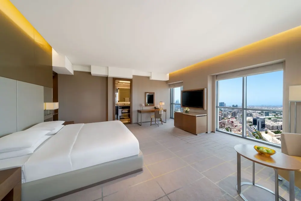A hotel bedroom with a large bed, a wall-mounted television, a desk, and floor-to-ceiling windows overlooking a city.