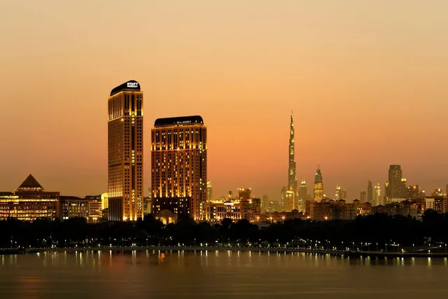 Dusk view across water of two illuminated Hyatt Regency towers with the Burj Khalifa and other city buildings lit behind them.