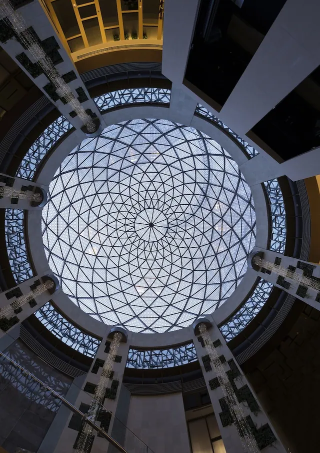 Looking up at a large geometric glass dome skylight in a multi-storey atrium, with columns decorated with greenery and hanging crystal strands.