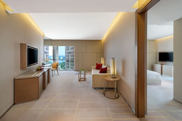 A suite living area with a cream sofa, round side table, wall-mounted television and a window looking onto high-rise buildings, with a bedroom and second television visible through the doorway on the right.