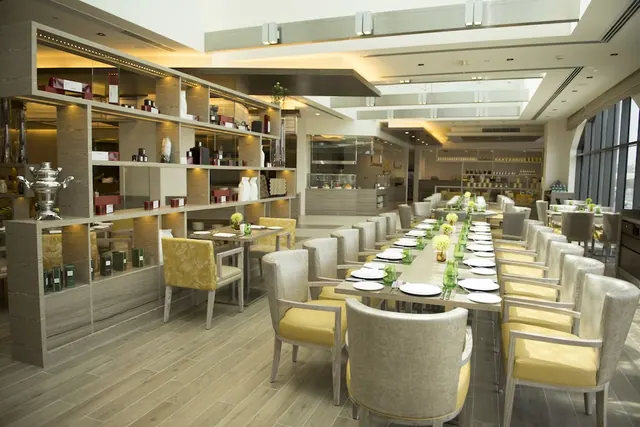 A restaurant dining room with long tables set with plates and green glasses, yellow-cushioned chairs, display shelving on the left, and a buffet counter in the background.