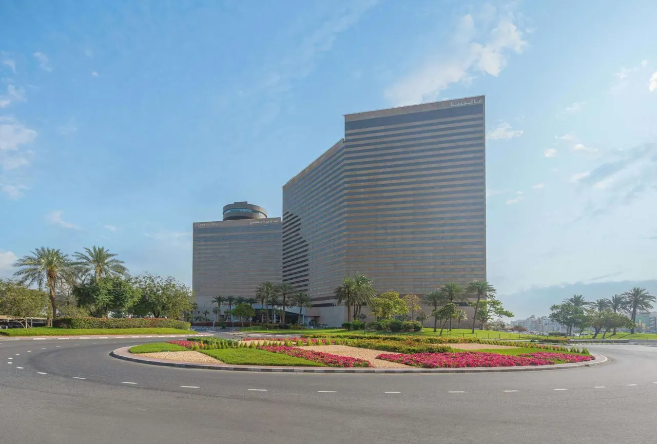 The exterior of a tall hotel tower stands behind a roundabout with flowerbeds, palm trees and a clear blue sky.