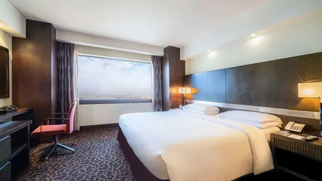 A made king bed with white linen sits across from a wide window with a hazy city view, next to a desk with a red chair and a bedside telephone.