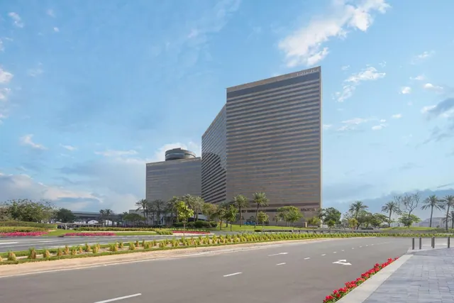 A brown-tinted high-rise hotel tower with a lower wing stands behind a wide road, a landscaped median and palm trees under a partly cloudy sky.