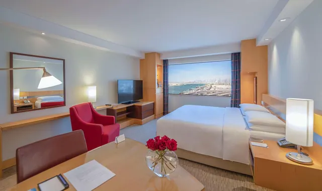 A hotel bedroom with a king bed, a red armchair, a wall-mounted television and a large window showing the sea and a city skyline.