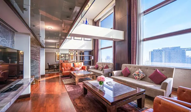 A double-height living area with a grey sofa set, orange leather armchairs, wooden coffee tables and large windows looking out over a city skyline.