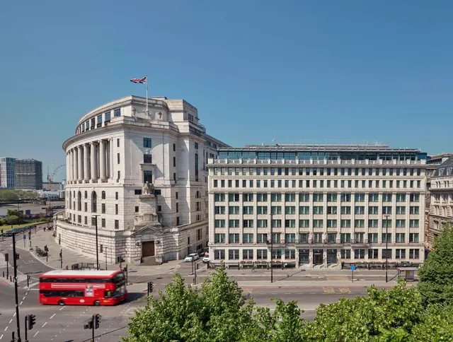 Exterior view of a large pale stone classical building with a Union flag on its roof, beside a long white stone office-style block, with a red double-decker bus on the street below.