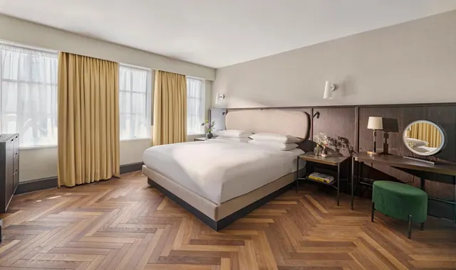 A bedroom with a king bed with white bedding, tall curtained windows, a herringbone wood floor, and a green stool beside a wall-mounted dressing table with a round mirror.