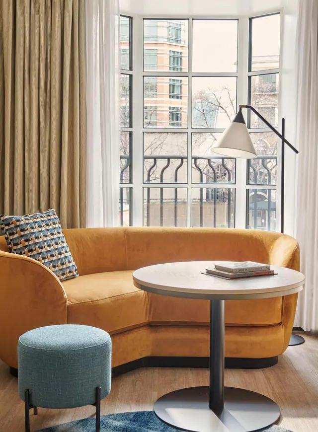 A seating area in a hotel room with a curved mustard velvet sofa, a round side table, a teal pouf and a bay window overlooking brick buildings.