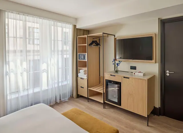 A hotel bedroom with a bed, an open wardrobe with hangers and towels, a safe, a mounted television and a glass-fronted minibar beside a window with sheer curtains that looks onto a neighbouring building.