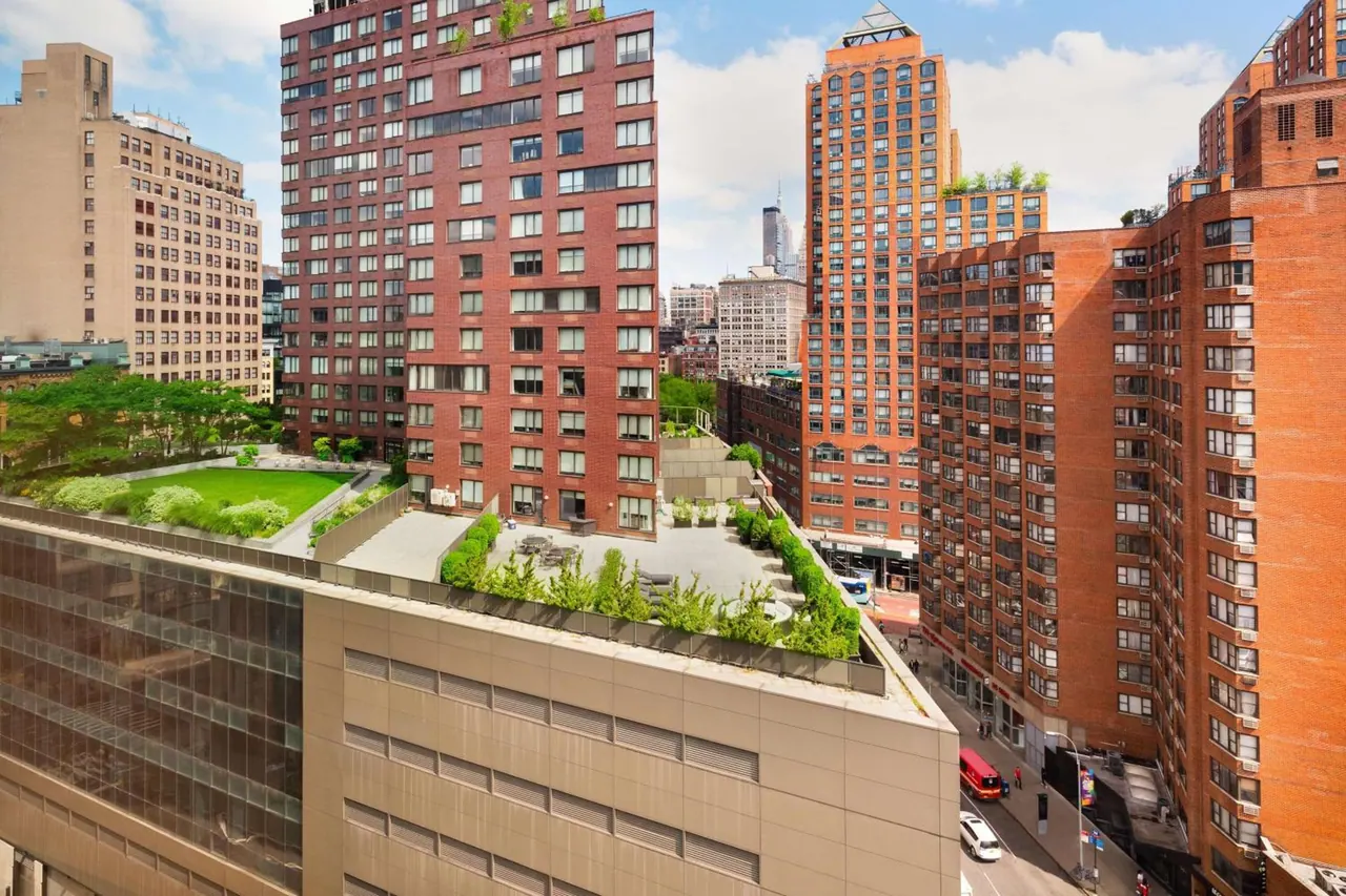 Elevated view over a rooftop garden and terrace toward brick high-rise buildings, with a street and parked vehicles below.