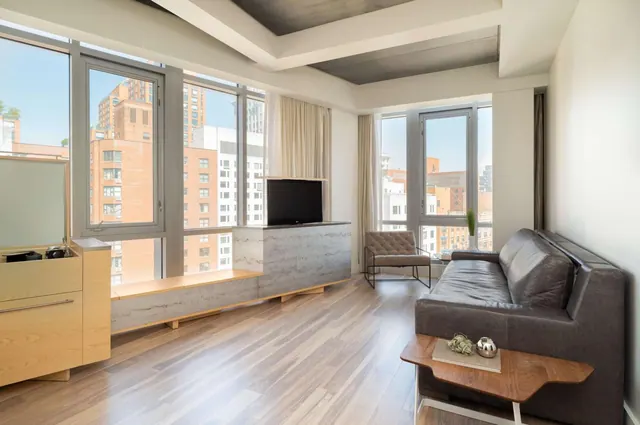 A living area with a grey leather sofa, a television on a stone-clad platform, and floor-to-ceiling windows looking onto city buildings.