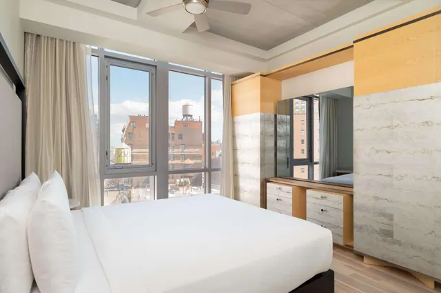 A bedroom with a made king bed, a work desk with drawers, a wardrobe, and floor-to-ceiling windows looking out over brick buildings and a water tower.