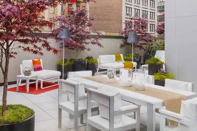 A rooftop terrace with white lounge seating, a dining table with a burlap runner, and Japanese maple trees in dark planters, with brick and stone buildings behind.