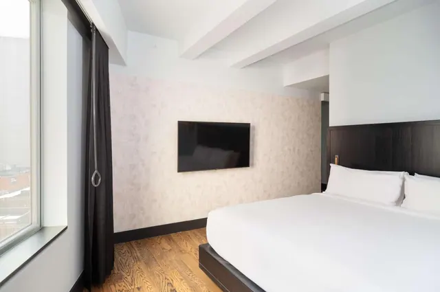 A bedroom with a king bed with white linens, a wall-mounted flat-screen television, and a large window with a dark curtain on the left.