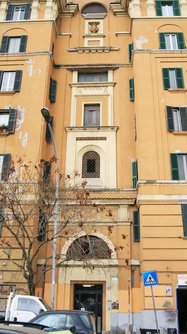 Exterior of a yellow-ochre apartment building with green shutters, a bare tree and a street lamp in front, and parked cars at the bottom edge.