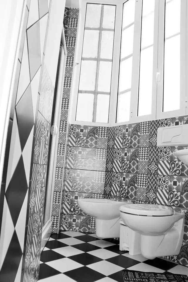 A black and white photo of a bathroom with patterned tiled walls, a checkerboard tiled floor, a wall-hung bidet and toilet, and a window with frosted panes above.