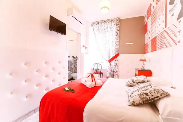 A bedroom with a red throw on a double bed, a wall-mounted flat-screen TV, a wall air conditioner, and a bathroom visible through an open door.