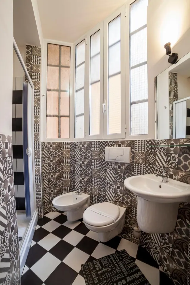A bathroom with patterned grey tiles, a black and white checkered floor, a wall-mounted bidet and toilet, a pedestal sink, and a row of frosted windows.