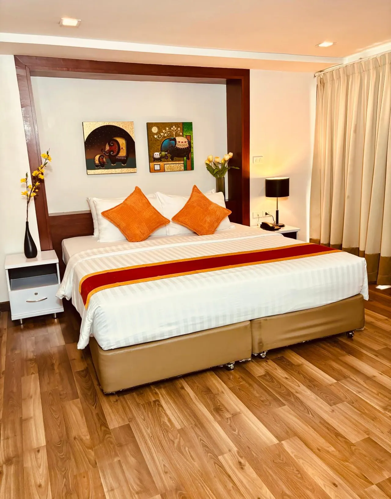 A hotel bedroom with a king bed, orange cushions, a red and orange bed runner, two framed paintings above the headboard and a wood-effect floor.