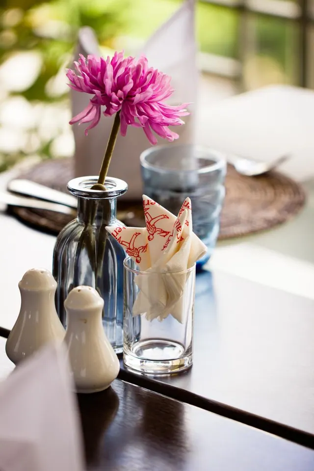 A pink flower stands in a glass bud vase beside white salt and pepper shakers, a folded napkin in a glass and a blue tumbler on a dining table.