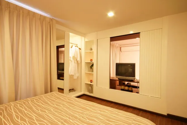 A bedroom with a patterned bed in the foreground, a wardrobe holding white robes, and shelves with candles, with an opening onto an adjacent room containing a television and a wall-mounted air conditioner.